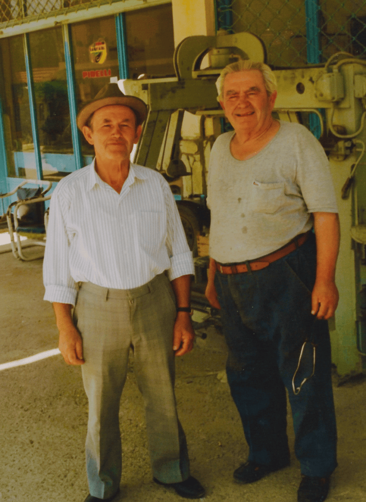 Photo restoration of a vintage film photograph. Two elderly men standing together outdoors in front of industrial machinery and a building with teal/turquoise window frames with a Pirelli sign visible. The man on the left is shorter, wearing a fedora hat and a light striped button-up shirt with grey trousers. The man on the right is taller with white hair, wearing a light grey t-shirt and dark jeans with a brown belt. Restore the faded old photo: correct the yellow/warm color cast typical of aged film photos, boost contrast and clarity, improve natural skin tones, sharpen facial details, brighten shadows while retaining a natural look. Well-preserved, naturally restored photograph with authentic character.